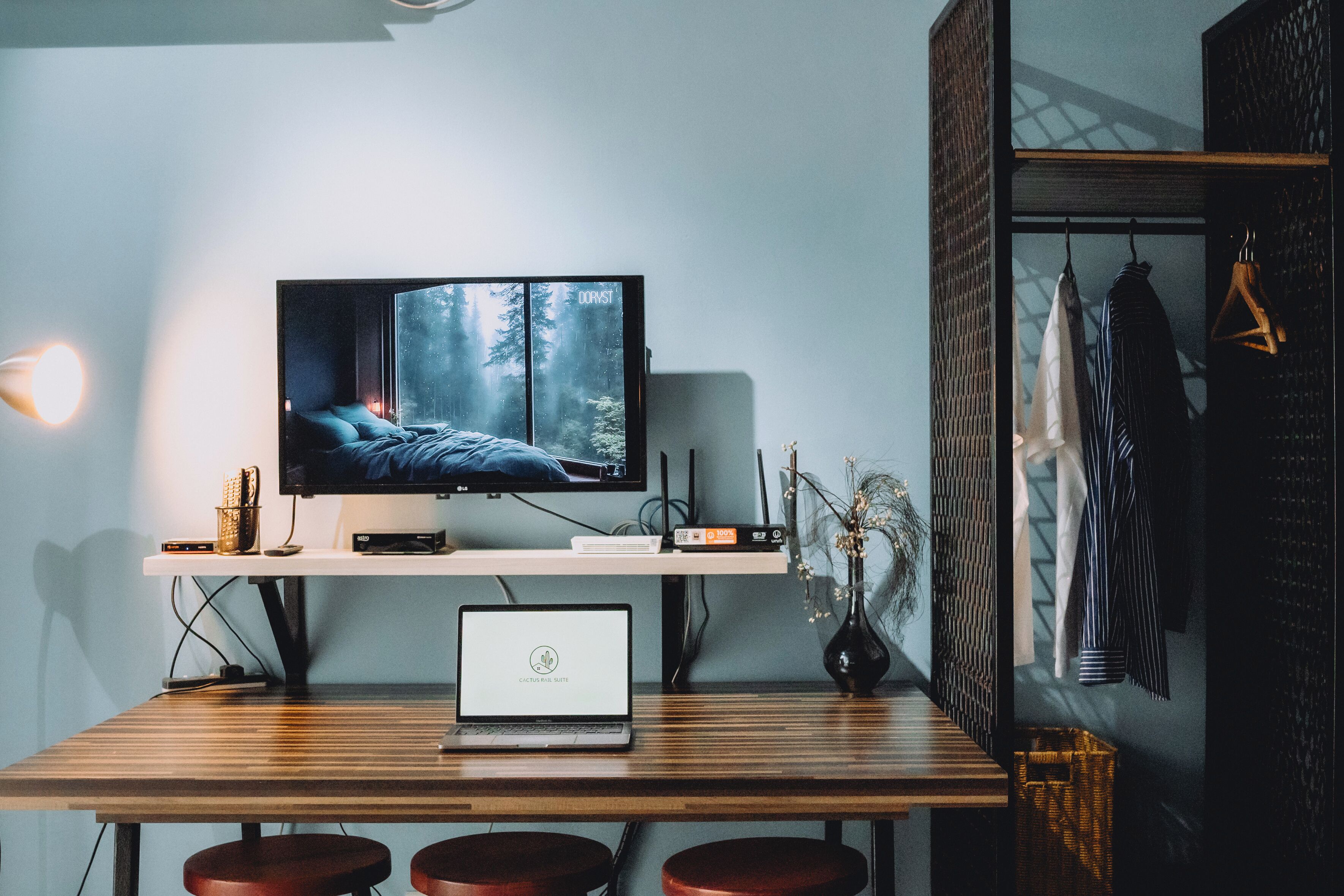 A modern room with a TV on a wooden shelf, a laptop on a table, and a clothing rack with hanging clothes.