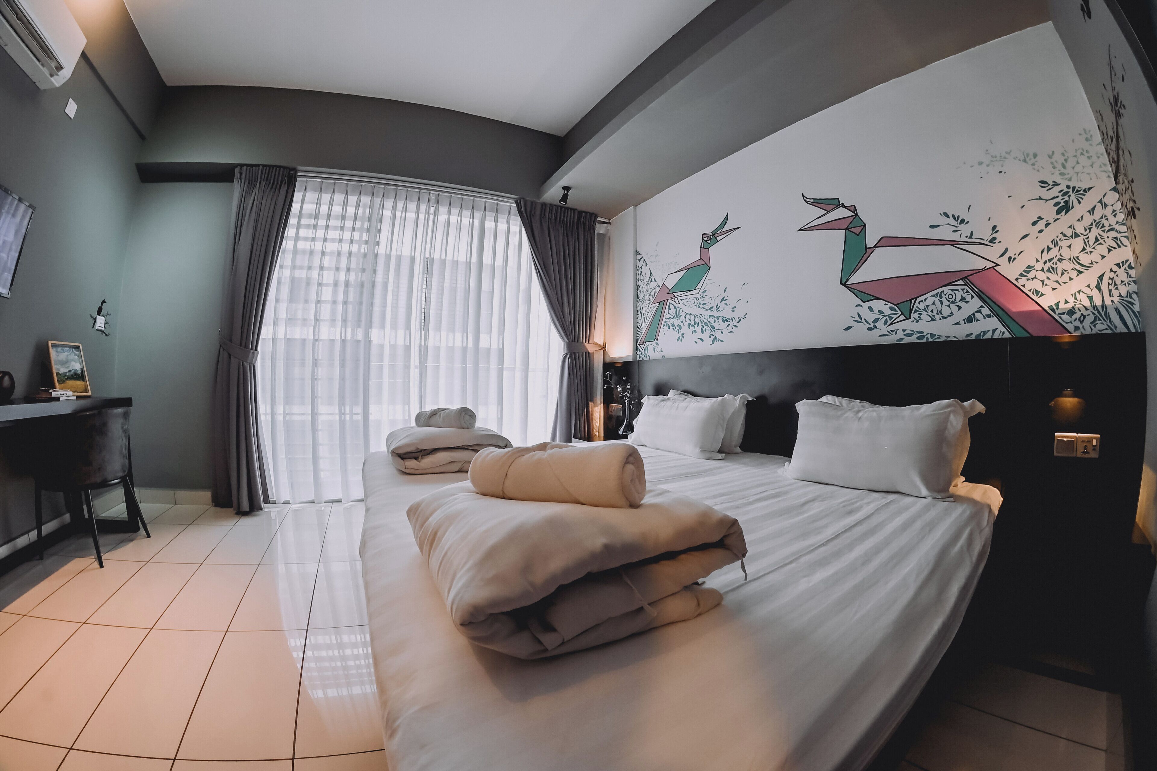 A modern hotel room with a large bed, a desk, and a view of the city through sheer curtains.