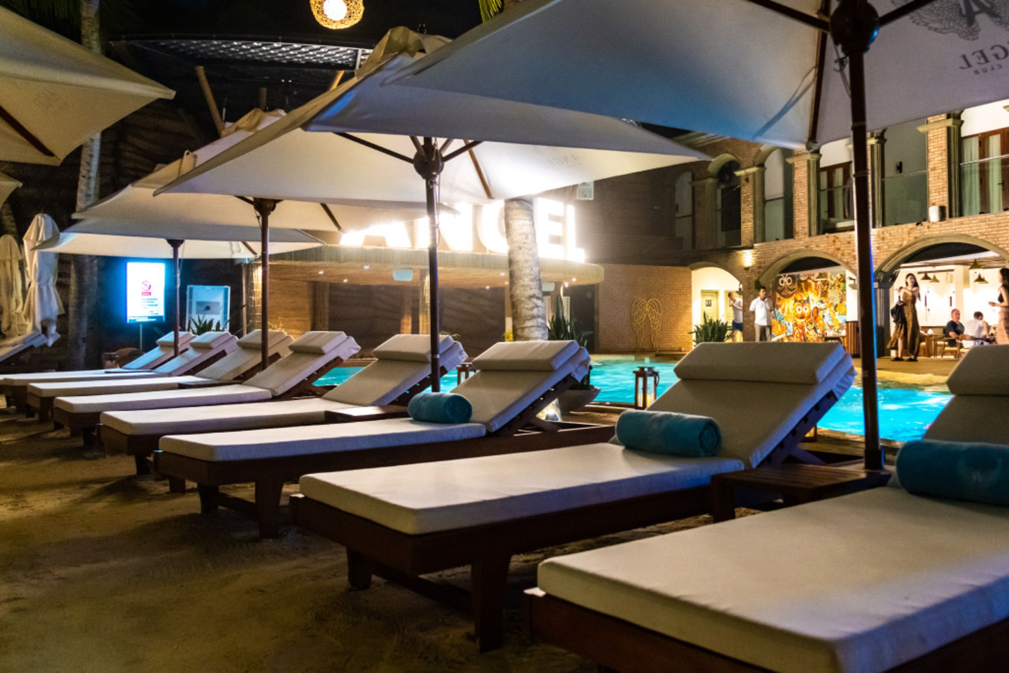 A poolside area with sun loungers and umbrellas, a brick building in the background, and a visible pool.