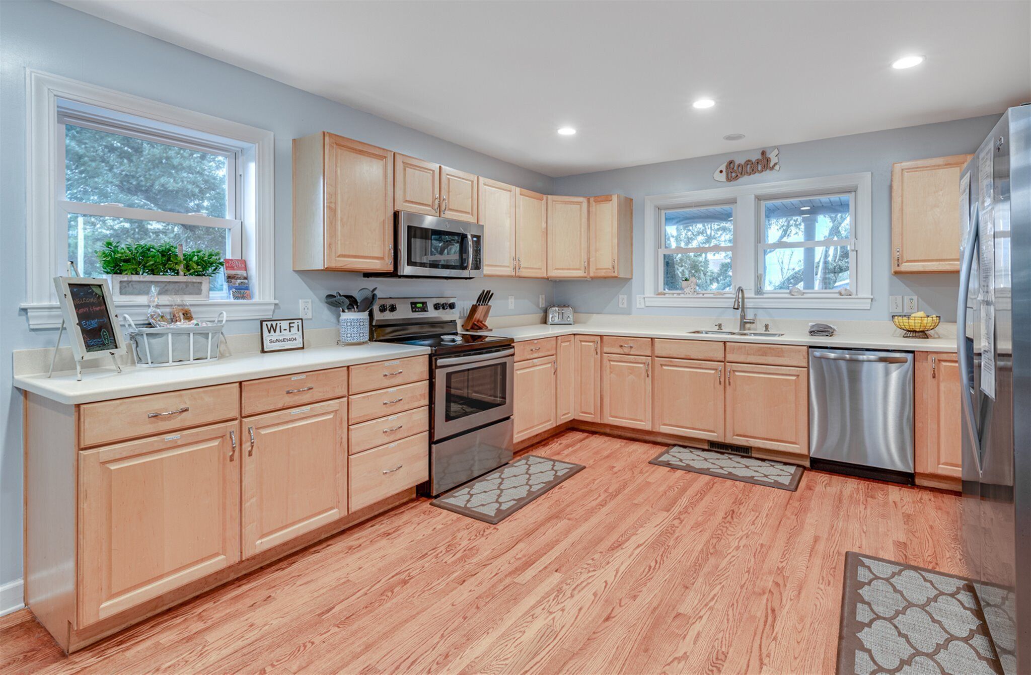 A modern kitchen with light wood cabinets, stainless steel appliances, and a view of the outdoors through large windows.