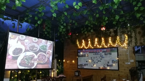 Un restaurante con piso a cuadros, plantas colgantes, una pantalla proyectando comida y una pizarra con el menú.