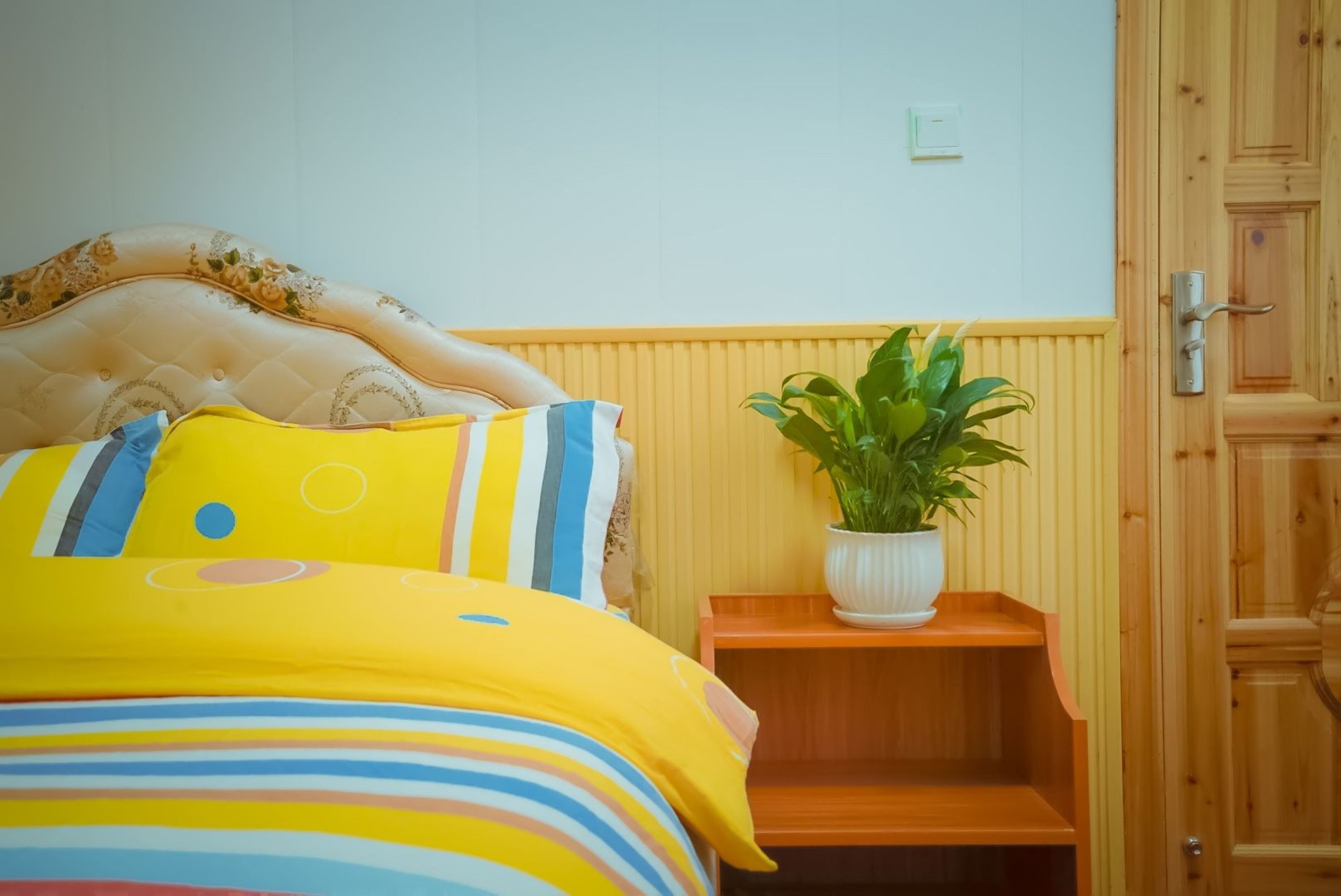 Een bed met een bloemrijk hoofdeinde, een geel gestreept dekbed en een kleine witte plant op een houten nachtkastje.