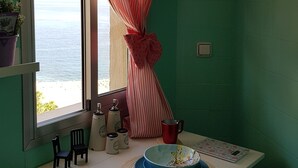 Une pièce avec une fenêtre, une table sur laquelle se trouvent un bol et des assiettes, deux tabourets, une horloge et une plante en pot.