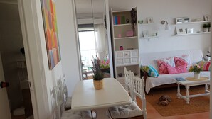 Un salon chaleureux avec un canapé blanc, des coussins colorés, une petite table sur laquelle est posée une plante, et une bibliothèque remplie de livres et d’objets décoratifs.