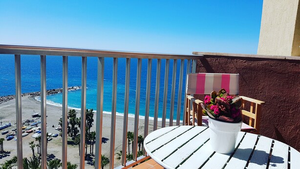 Un balcon avec vue sur la plage et l’océan, une table blanche avec une chaise à rayures et une plante en pot ornée de fleurs.