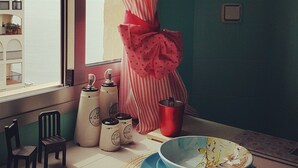 Une pièce avec une fenêtre, des rideaux à rayures, un ruban rouge, une chaise, une table avec une assiette bleue, une bouteille blanche et un pichet en métal.