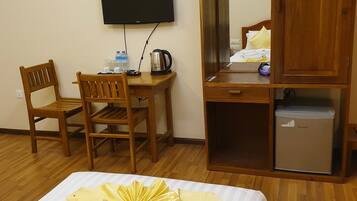 Quarto de hotel com cama, escrivaninha e cadeira, televisão, guarda-roupa e um cesto de lixo pequeno.