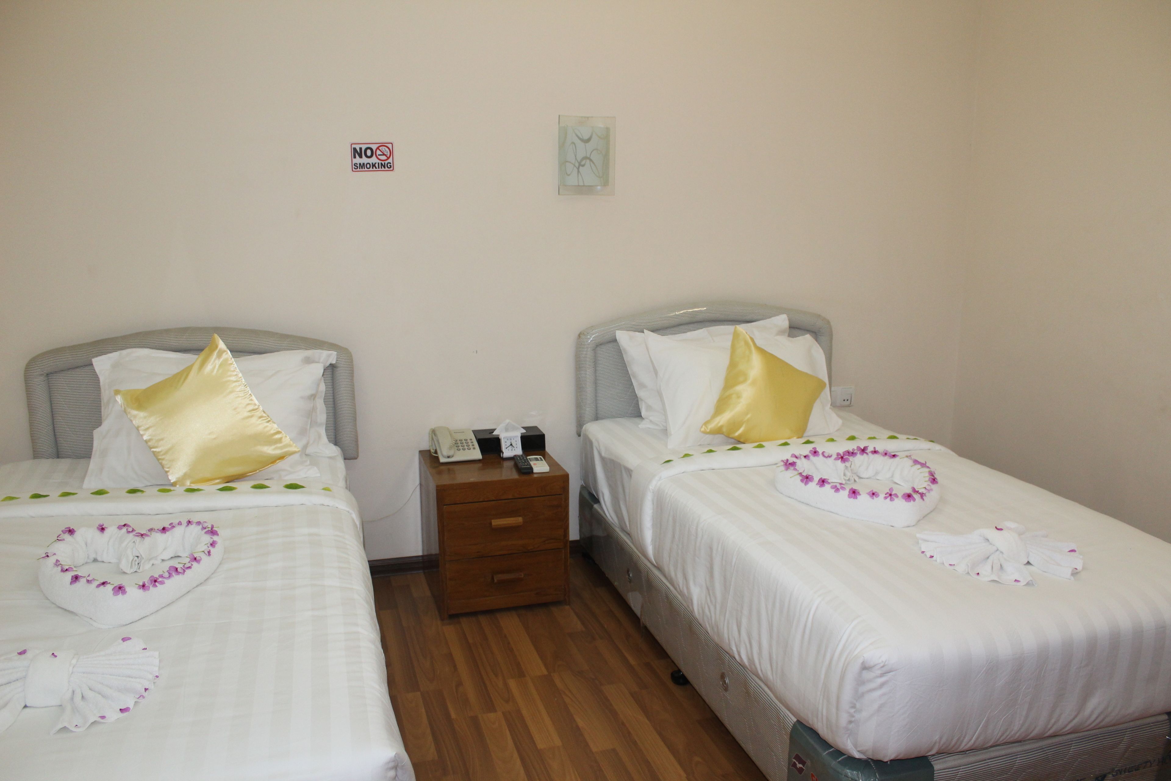 A hotel room with two beds, each with white bedding and decorative pillows. There is a wooden nightstand between the beds with a telephone and a small box. A 'No Smoking' sign is visible on the wall.