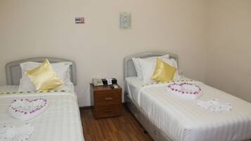 A hotel room with two beds, each with white bedding and decorative pillows. There is a wooden nightstand between the beds with a telephone and a small box. A 'No Smoking' sign is visible on the wall.