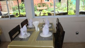 A dining area with tables set for four, each with a white tablecloth, silverware, and folded napkins. Large windows offer a view of a garden and a brick building. - Law Ka Thit Sar Hotel - Hostel (Nyaung-U)