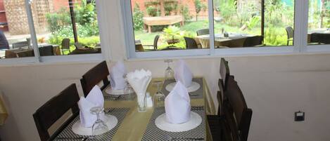 Comedor con mesas dispuestas para cuatro, cada una con mantel blanco, cubiertos y servilletas dobladas. Amplias ventanas ofrecen vistas a un jardín y un edificio de ladrillo.