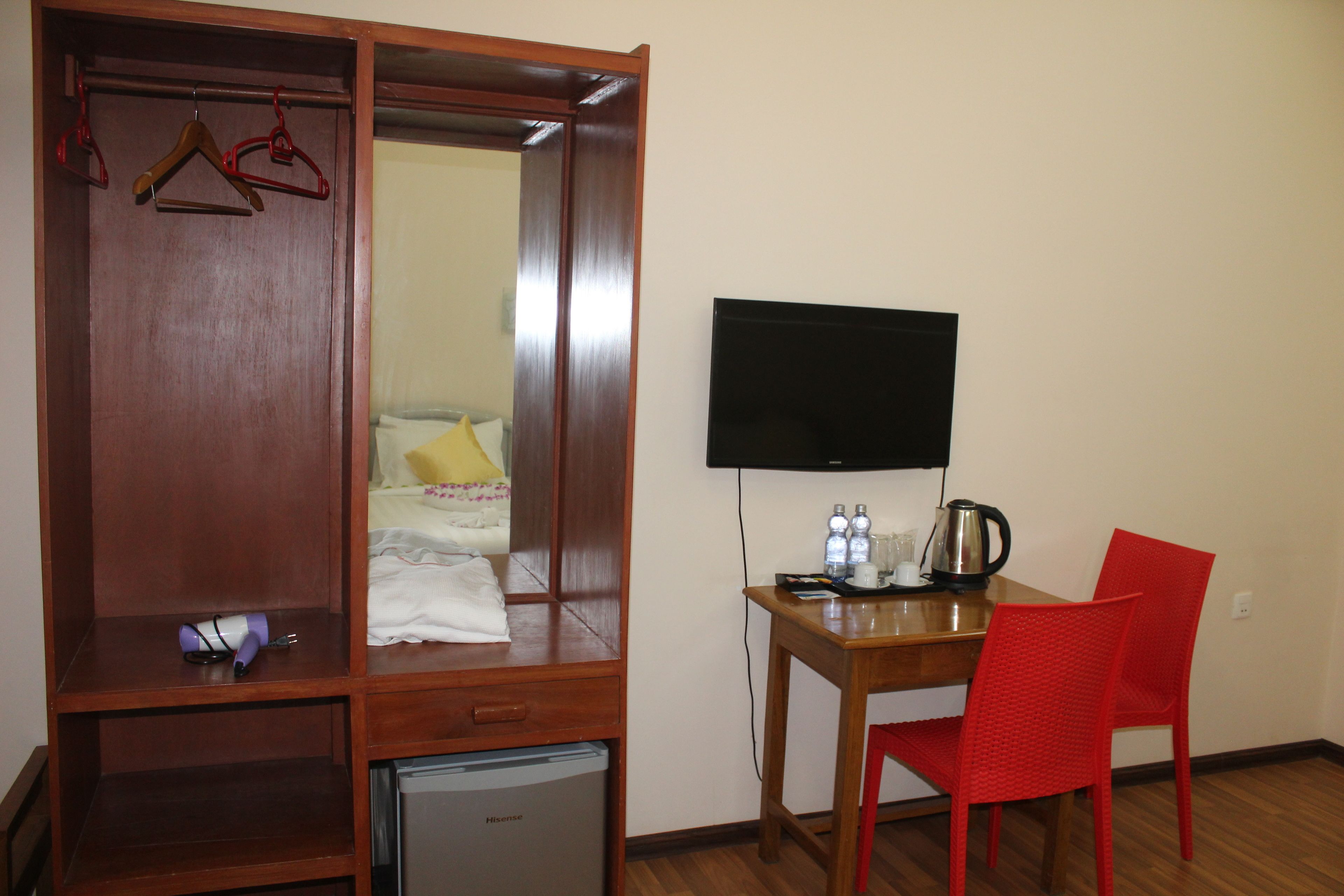 Une chambre compacte comprenant une armoire en bois, un petit réfrigérateur, un bureau avec une chaise, un téléviseur à écran plat et une bouilloire.