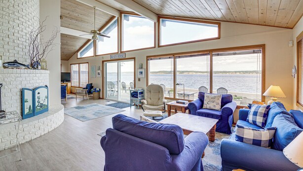 A spacious living room with a view of the sea, featuring a fireplace, comfortable seating, and a ceiling fan. - Beachfront Whidbey Island Home + Apartment! (Langley)