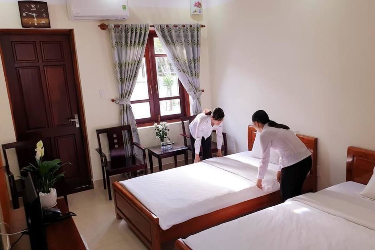 A hotel room with two beds, a desk, a chair, a potted plant, and a window with curtains.