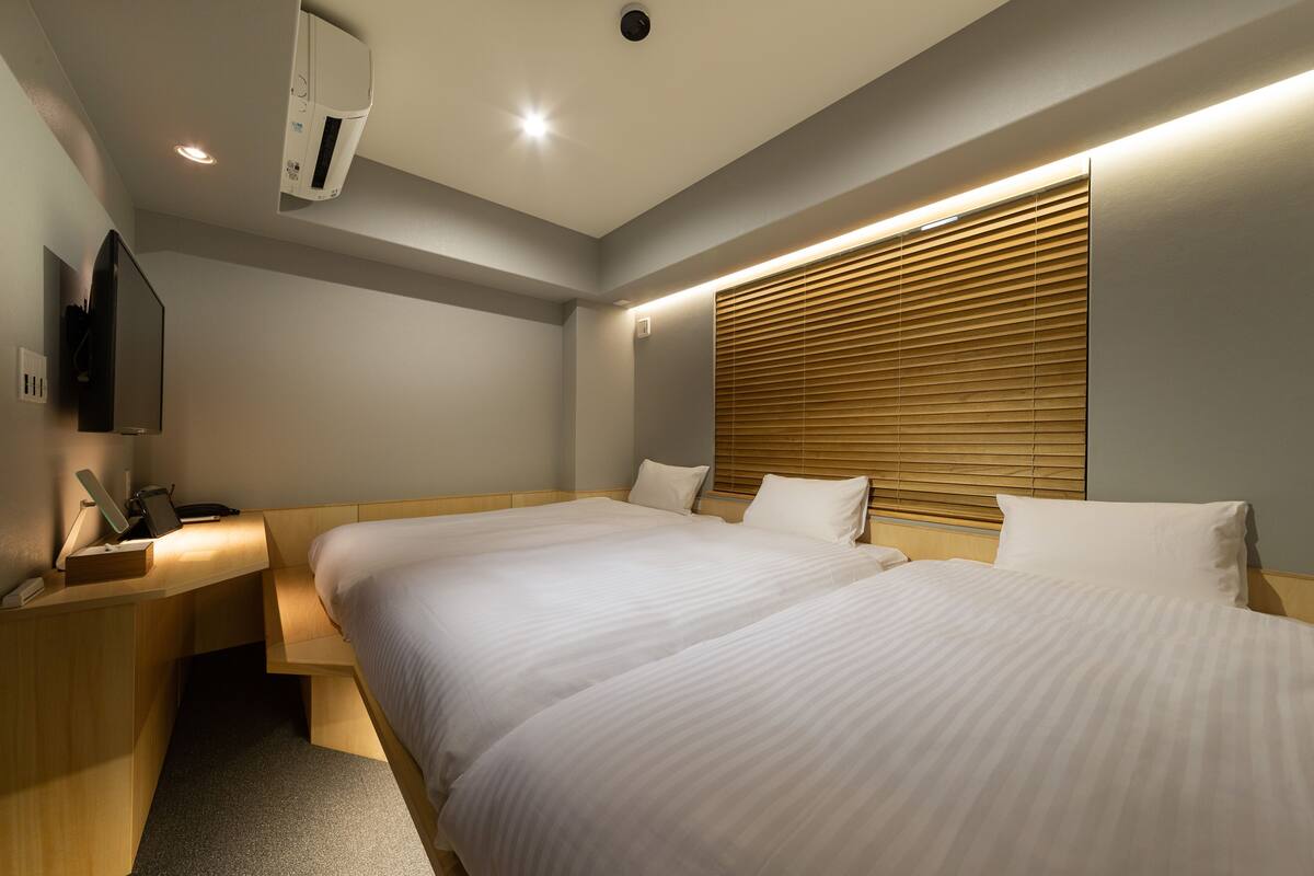 Ein Hotelzimmer mit zwei Betten, einem Schreibtisch mit Laptop, einem Flachbildfernseher und einem Fenster mit Jalousien.