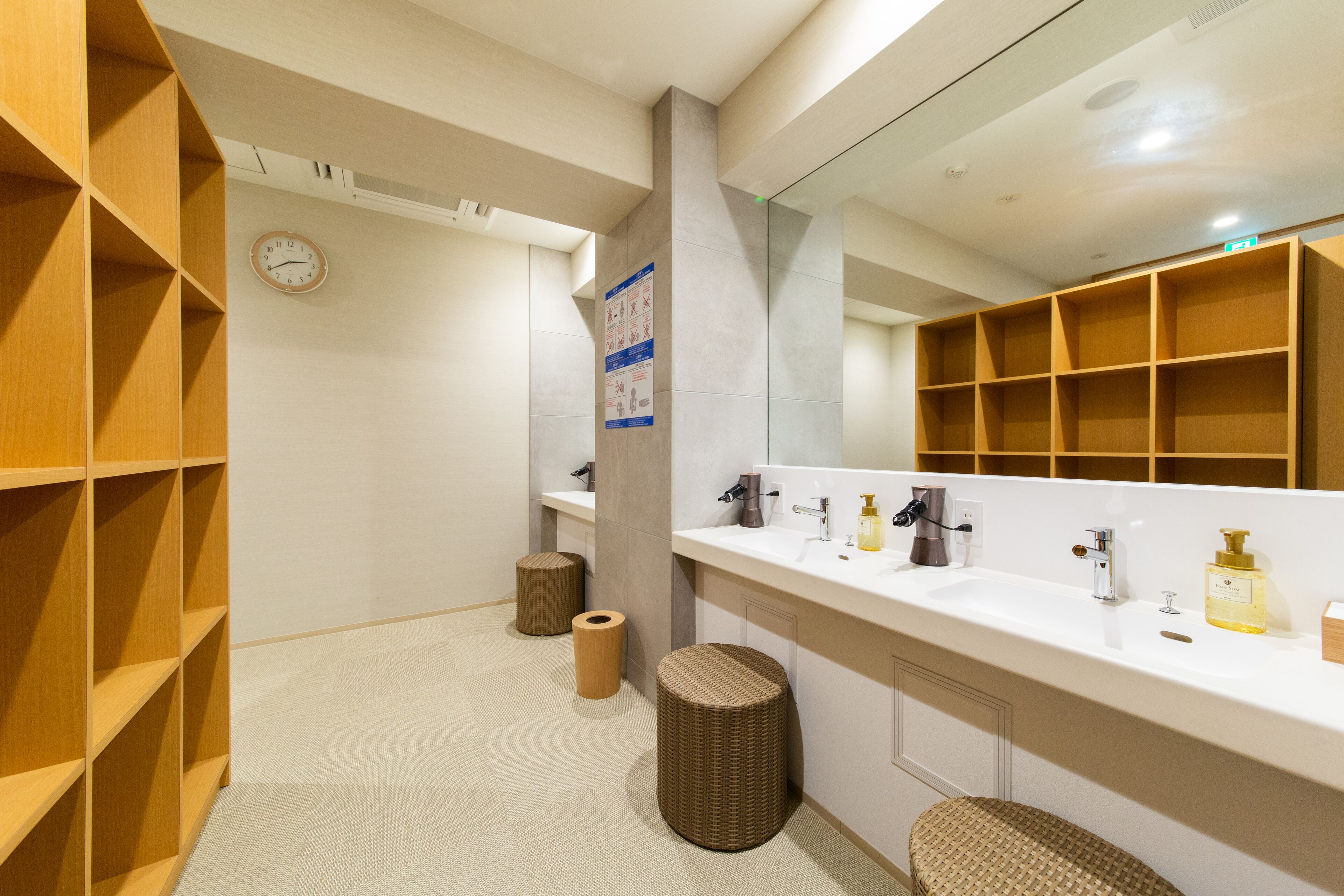 A modern bathroom with a white sink, mirror, and wooden shelves. There is a clock on the wall and a poster.