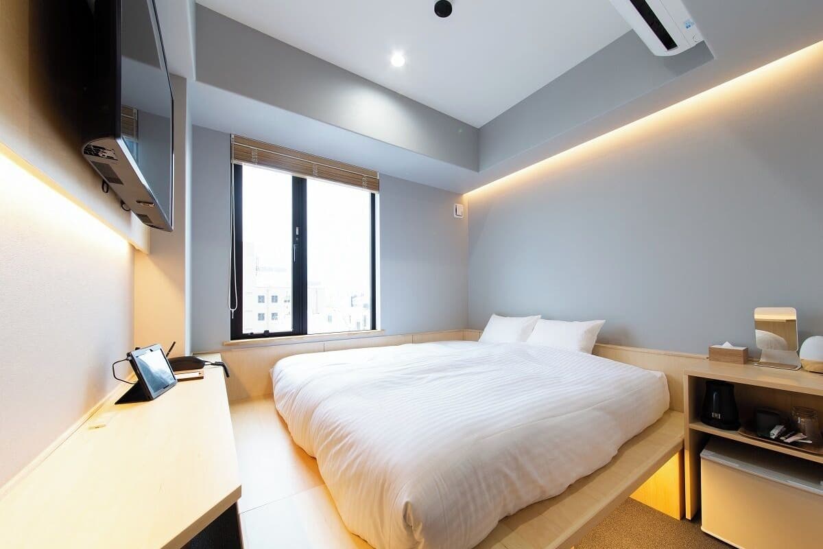 Ein modernes Hotelzimmer mit einem großen Bett, Nachttisch und Schreibtisch mit Stuhl. Es gibt ein Fenster mit Jalousien, einen flachbildfernseher an der Wand und eine Klimaanlage.