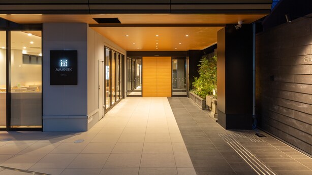 Una entrada de hotel moderna con un letrero que dice "AMANEK HOTEL", fachada de vidrio y un camino de ladrillos.