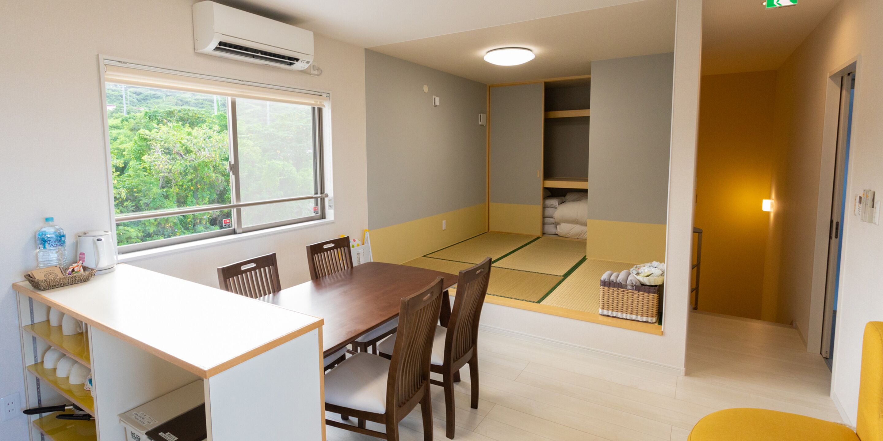 Une pièce comprenant une table à manger, des chaises, une fenêtre donnant sur un espace verdoyant, un coin avec un tatami et un petit placard.