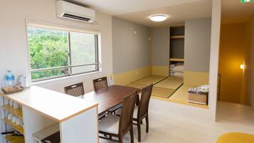 Une pièce comprenant une table à manger, des chaises, une fenêtre donnant sur un espace verdoyant, un coin avec un tatami et un petit placard.