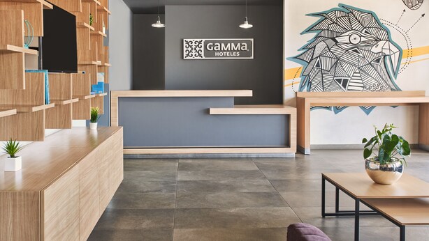 A modern hotel reception area with a wooden reception desk, a flat-screen TV, and a logo for Gamma Hotels. There is a large wall mural and a potted plant on the right. - Gamma Querétaro (Querétaro)
