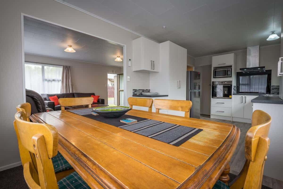 A dining area with a wooden table, chairs, and a bowl. There is a kitchen in the background with white cabinets and appliances.