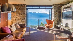 A modern living room with a stone wall, leather sofa, and a view of a lake and mountains. - Bellbrae by MajorDomo | Stylish 5-Bedroom Villa with Sauna, Spa Pool & Sweeping Lake Wakatipu Views (Queenstown)