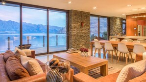 A modern living room with a large window overlooking a lake and mountains, a stone accent wall, and a dining area with a kitchen in view. - Bellbrae by MajorDomo | Stylish 5-Bedroom Villa with Sauna, Spa Pool & Sweeping Lake Wakatipu Views (Queenstown)