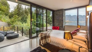 A modern bedroom with a large glass door leading to a balcony, a bed with a view of the outdoors, and a red chair. - Bellbrae by MajorDomo | Stylish 5-Bedroom Villa with Sauna, Spa Pool & Sweeping Lake Wakatipu Views (Queenstown)