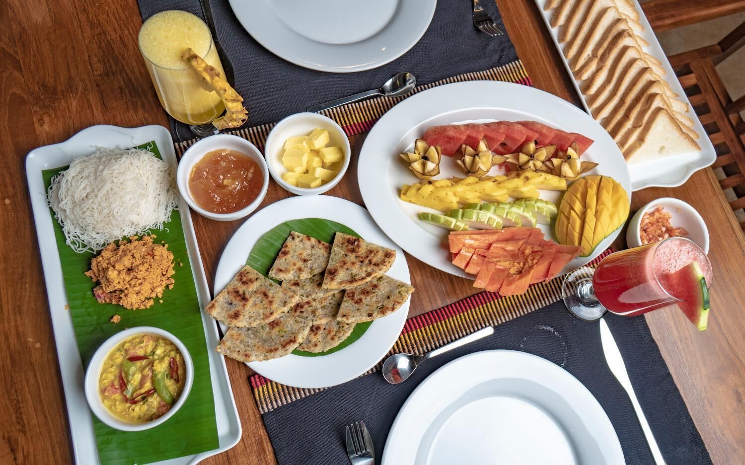 A table with various dishes including rice, curry, and grilled vegetables, along with a fruit salad and a drink.
