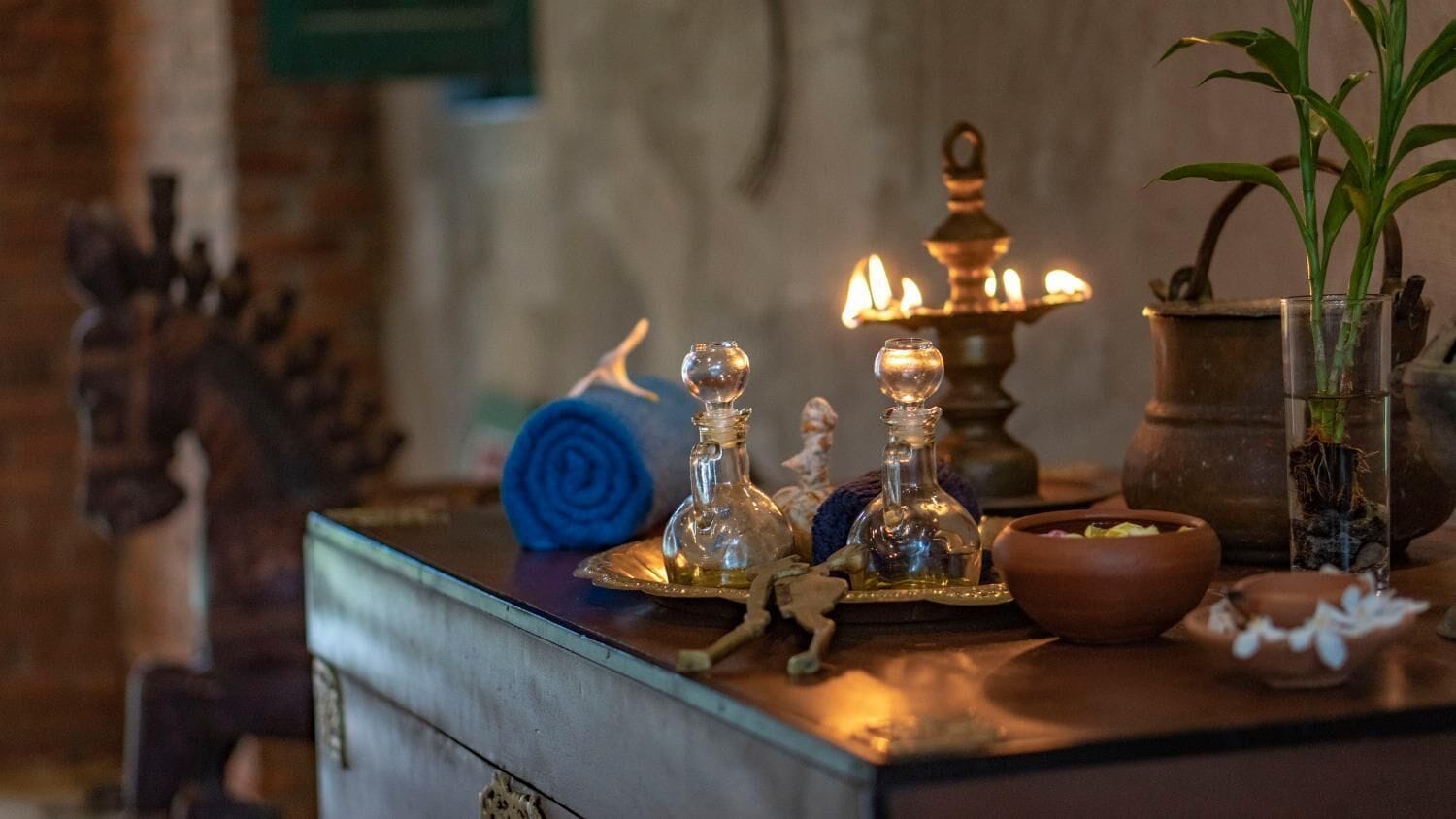 A table with a brass lamp, glass bottles, a blue rose, a small plant, and a clay pot.