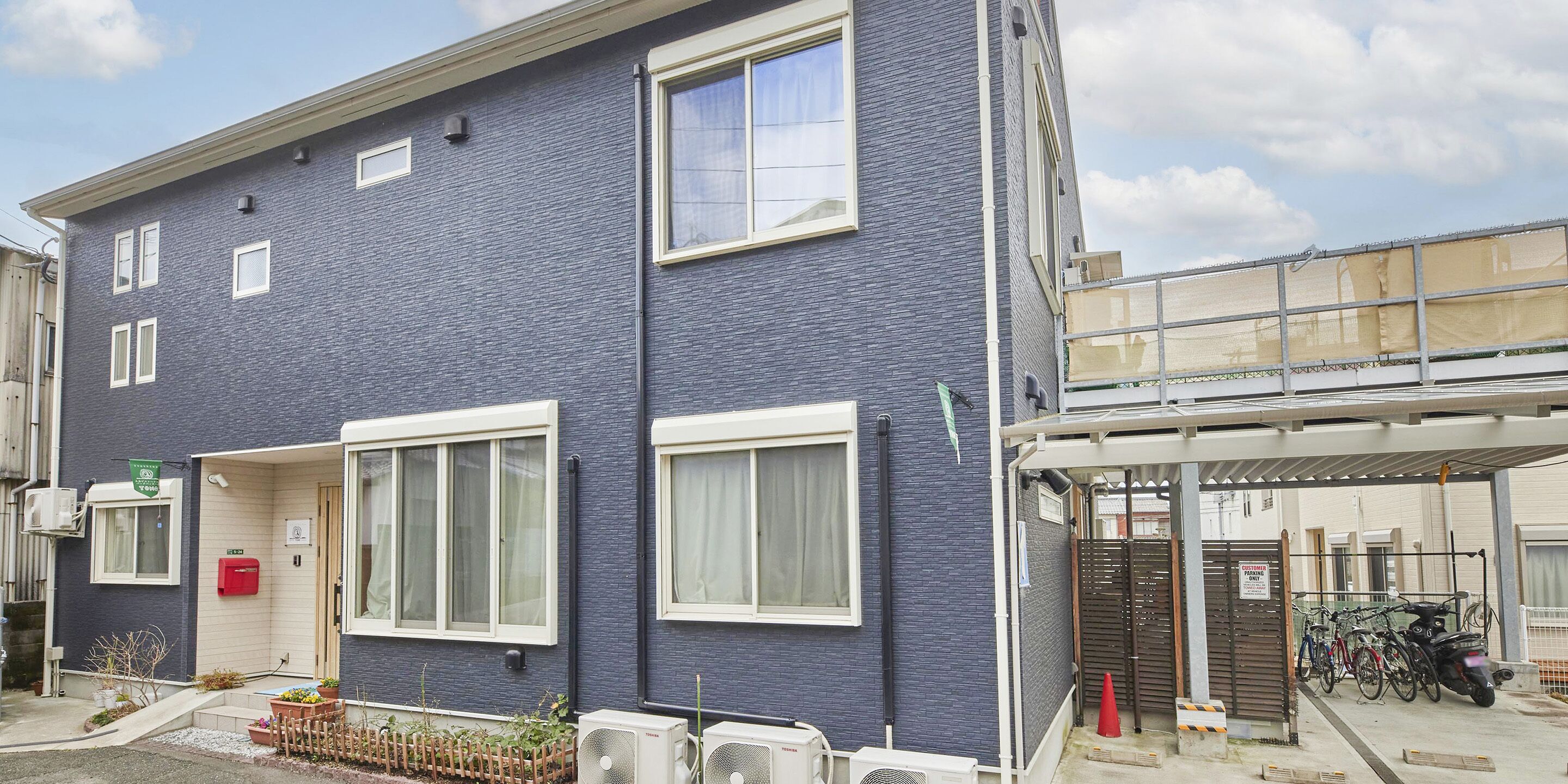 Un edificio de dos pisos con fachada azul oscuro, ventanas con marcos blancos y un área de entrada cubierta donde se estacionan bicicletas.