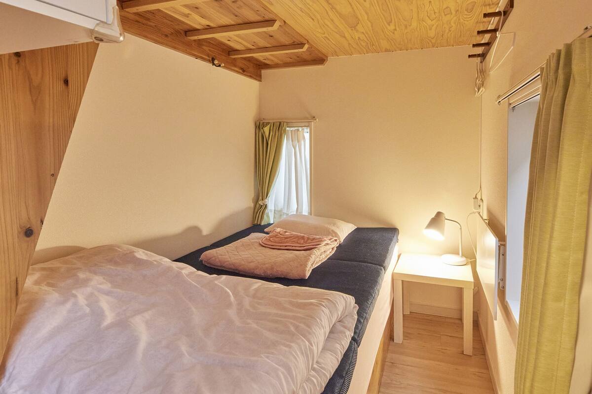 Een kleine, gezellige kamer met een bed, een nachtkastje met een lamp en een raam met gordijnen.