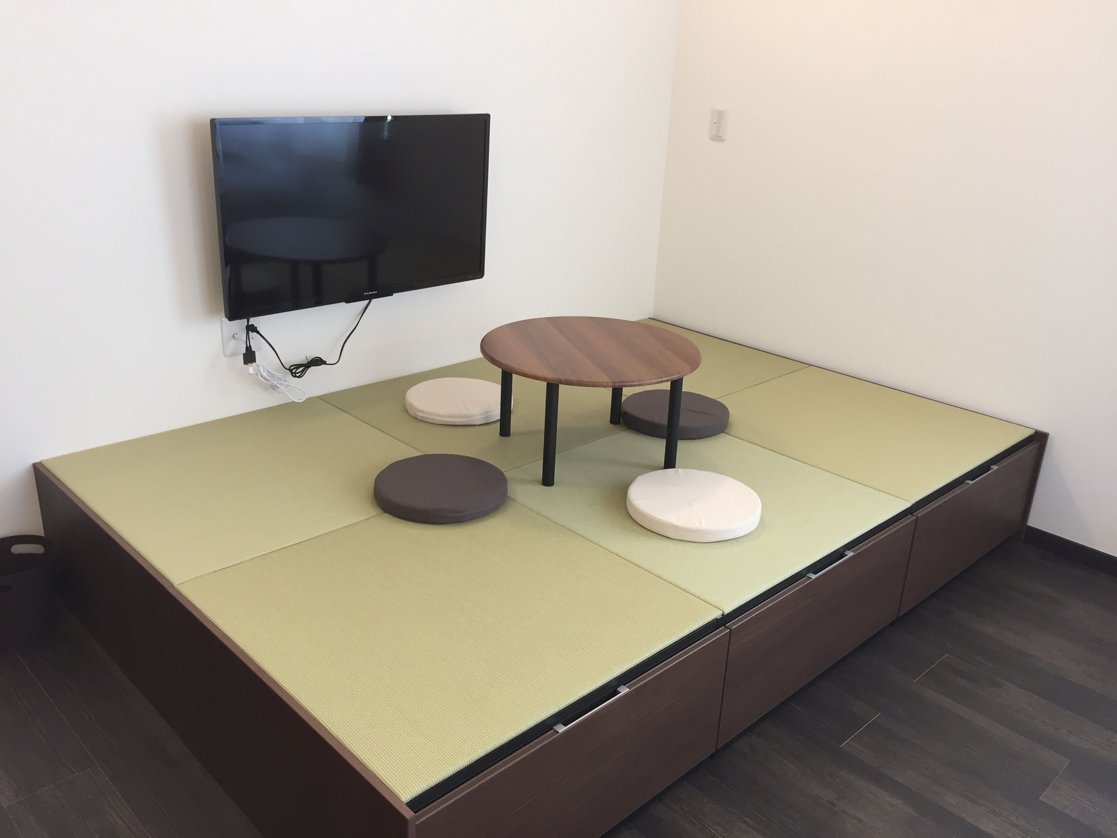A tatami mat room with a television mounted on the wall, a small round table, and two cushions.