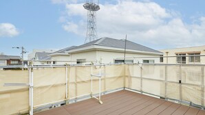 Una terraza en la azotea con cubierta de madera, cerca con malla blanca, y un edificio de techo gris al fondo.