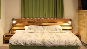 Ein hölzernes Bett mit floralem Überwurf und zwei Kissen, ein grüner Vorhang und eine Holzkommode.