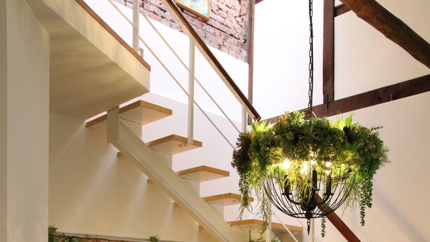 Ein modernes Wohnzimmer mit einem braunen Ecksofa, einem kleinen weißen Beistelltisch, einer schwarzen Stehlampe, einem Hängeleuchter mit Pflanzen und einer Treppe, sowie einer Ziegelwand mit einem gerahmten Gemälde.