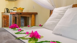 Una cama con almohadas blancas y dosel, una mesita de noche de madera con cesta de frutas, y una kitchenette al fondo.