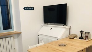 Un espacio habitable compacto con una mesa de comedor de madera, sillas blancas, un televisor sobre un mueble bajo, una pequeña kitchenette y un letrero que dice "BOUT DU MONDE".