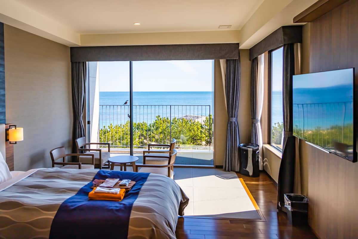 Ett hotellrum med en säng, ett skrivbord, en stol, en TV och en balkong med utsikt över havet.