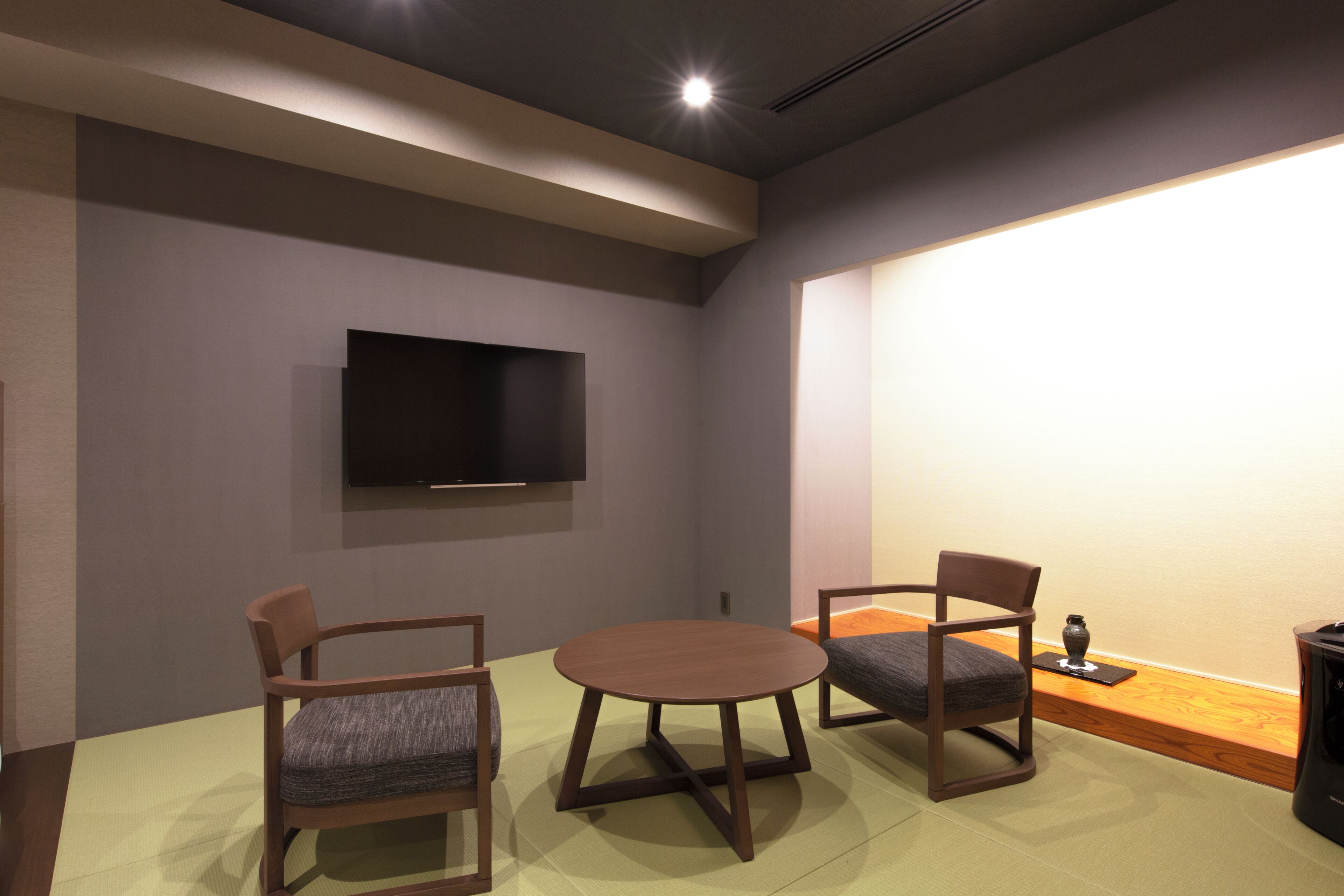 Une pièce dotée d’un téléviseur à écran plat fixé au mur, de deux chaises en bois avec des coussins, d’une table ronde et d’un petit vase décoratif posé sur le sol.