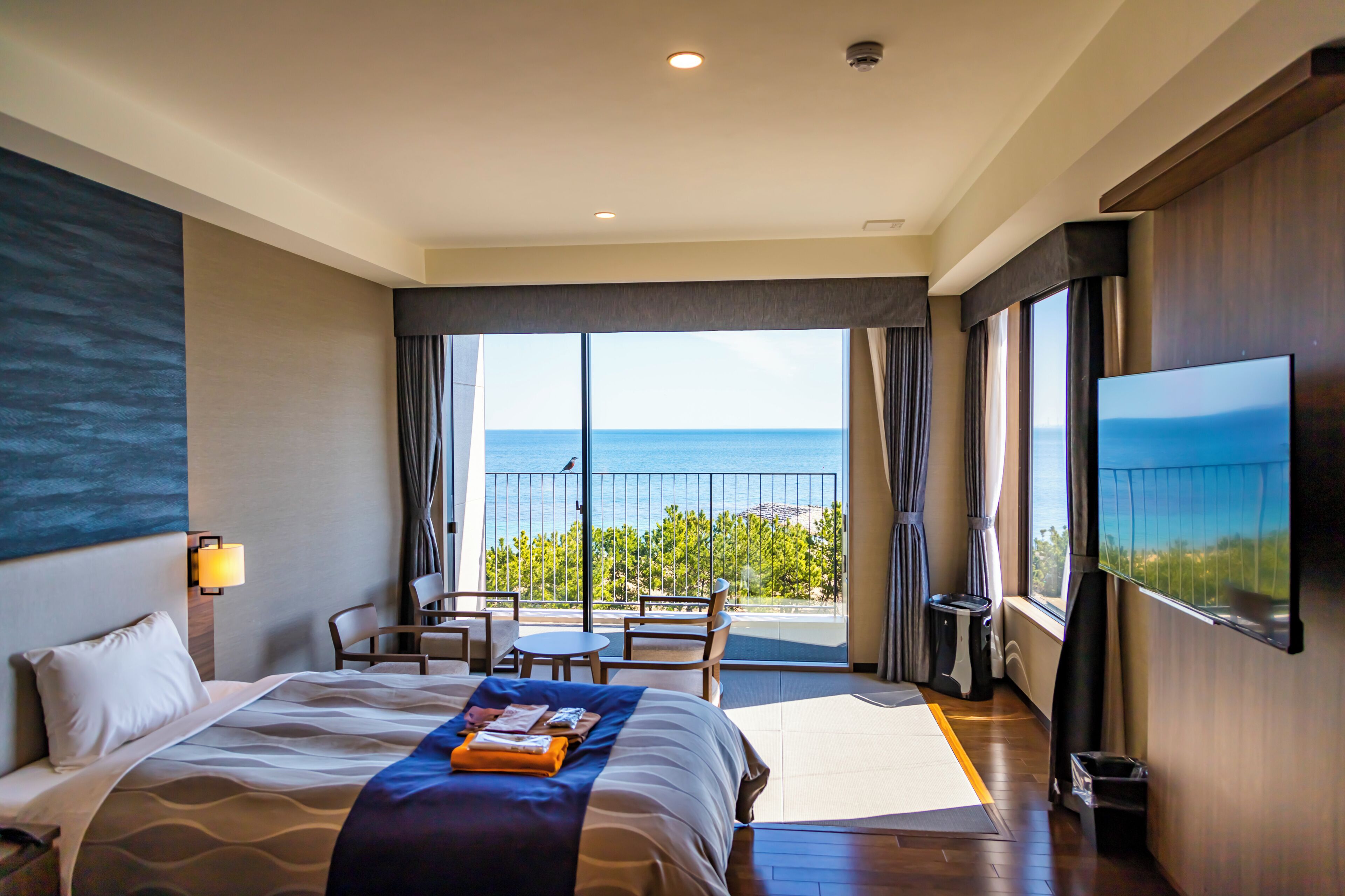 A hotel room with a bed, a TV, a balcony with a view of the sea, and a table with chairs.