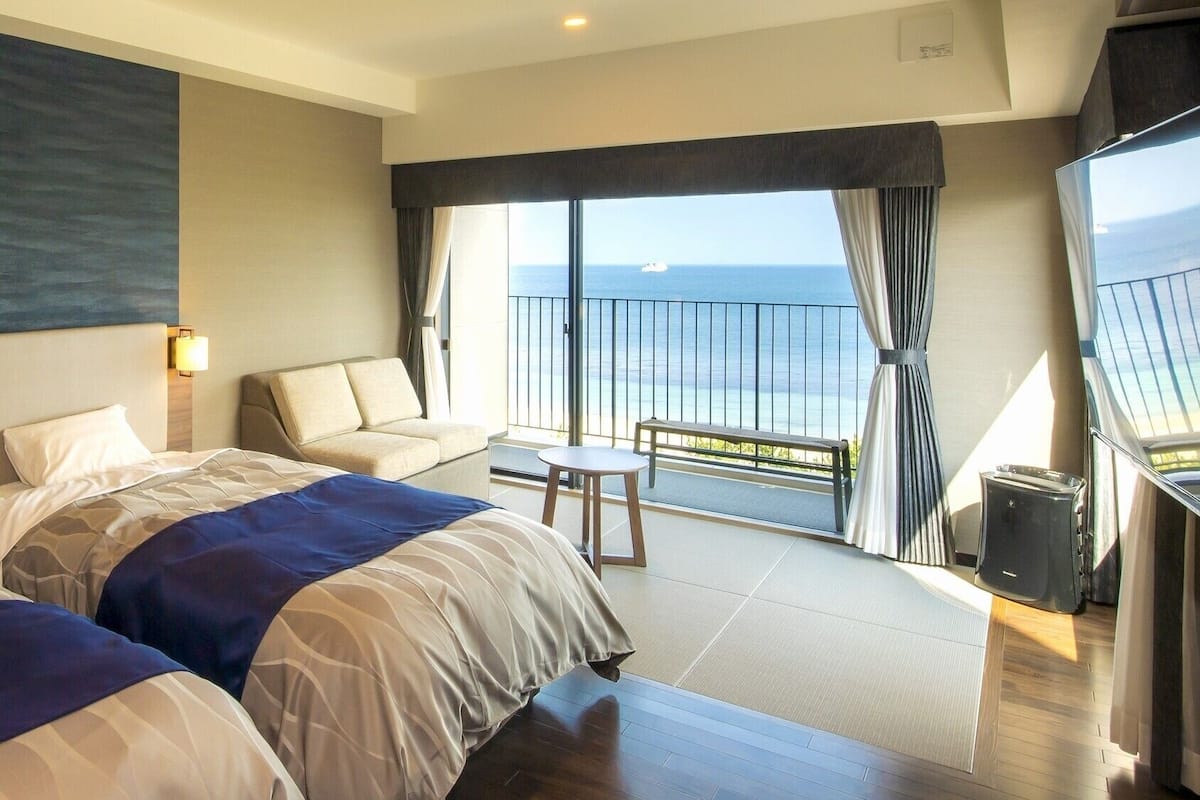 A hotel room with two beds, a sofa, a small table, and a balcony with a view of the sea.