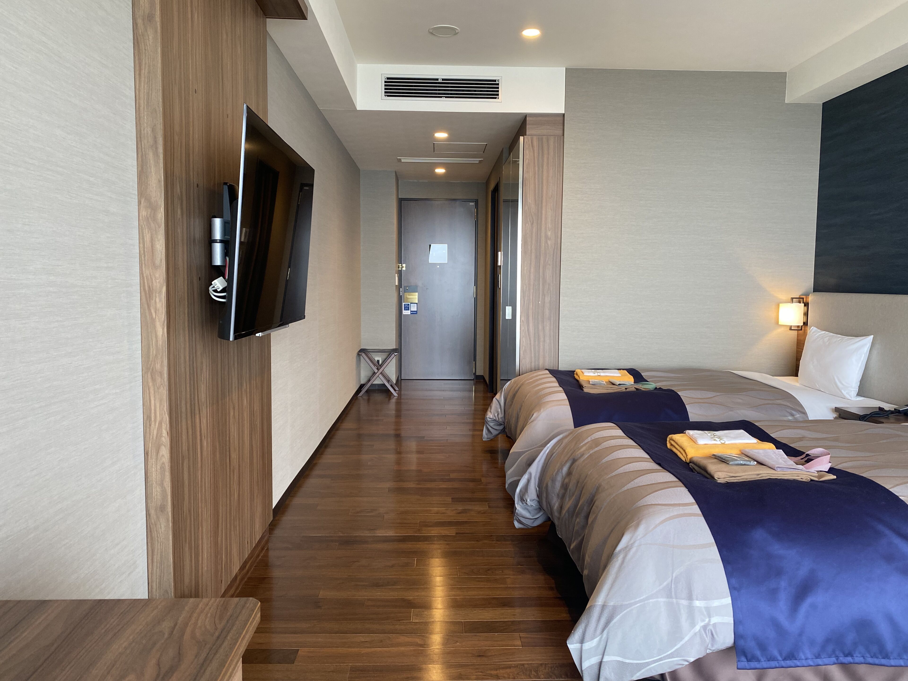 Quarto de hotel com piso de madeira, televisão na parede, cama com manta azul e mesa de cabeceira com abajur.