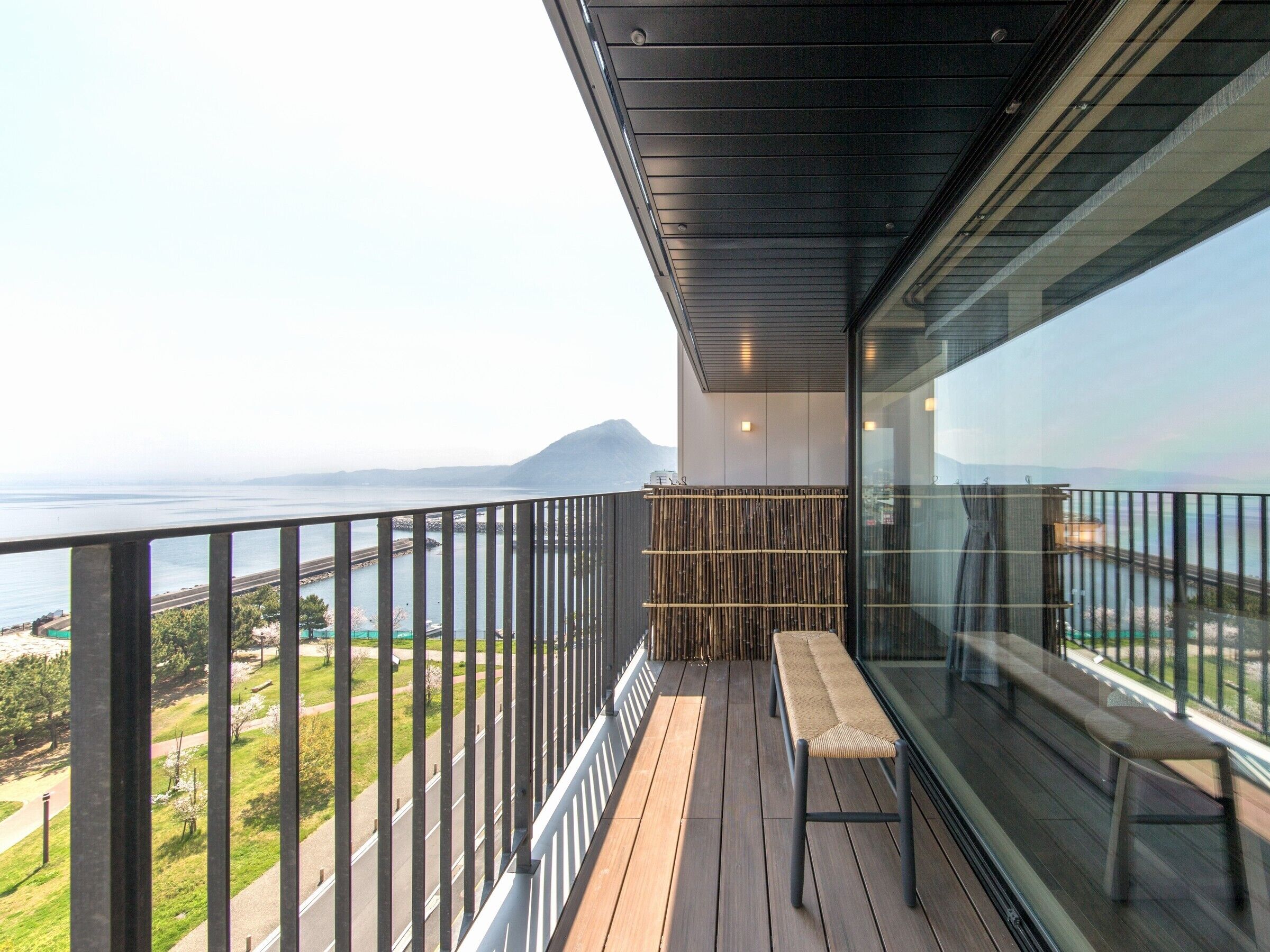 A balcony with a view of the sea and mountains, a wooden floor, and a bench.