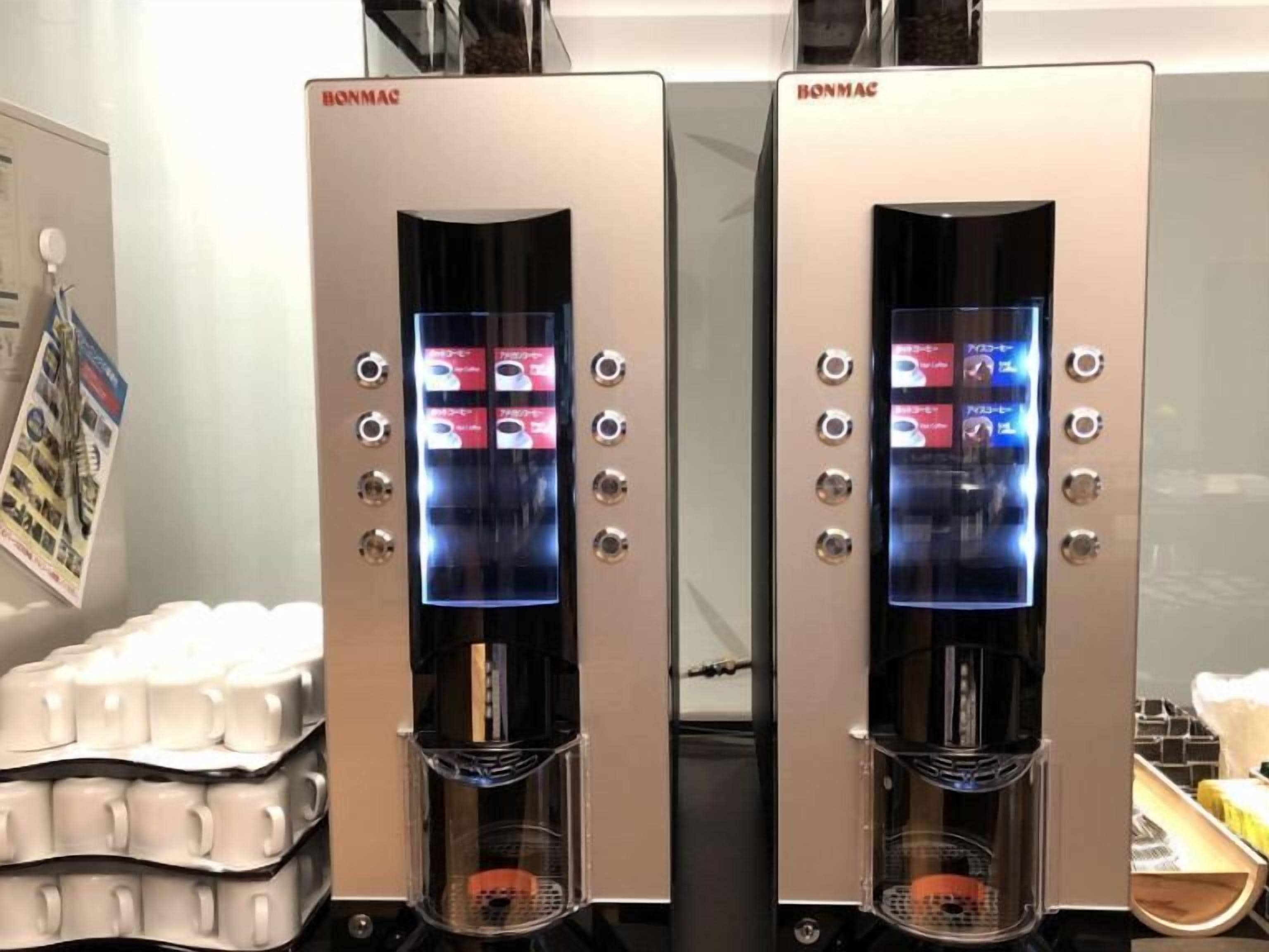 Dos máquinas de café con pantallas digitales, una pila de tazas blancas y un cartel en la pared.