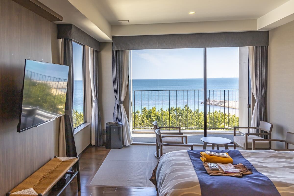 A hotel room with a large bed, a TV mounted on the wall, a balcony with a view of the sea, and a small table and chair set.
