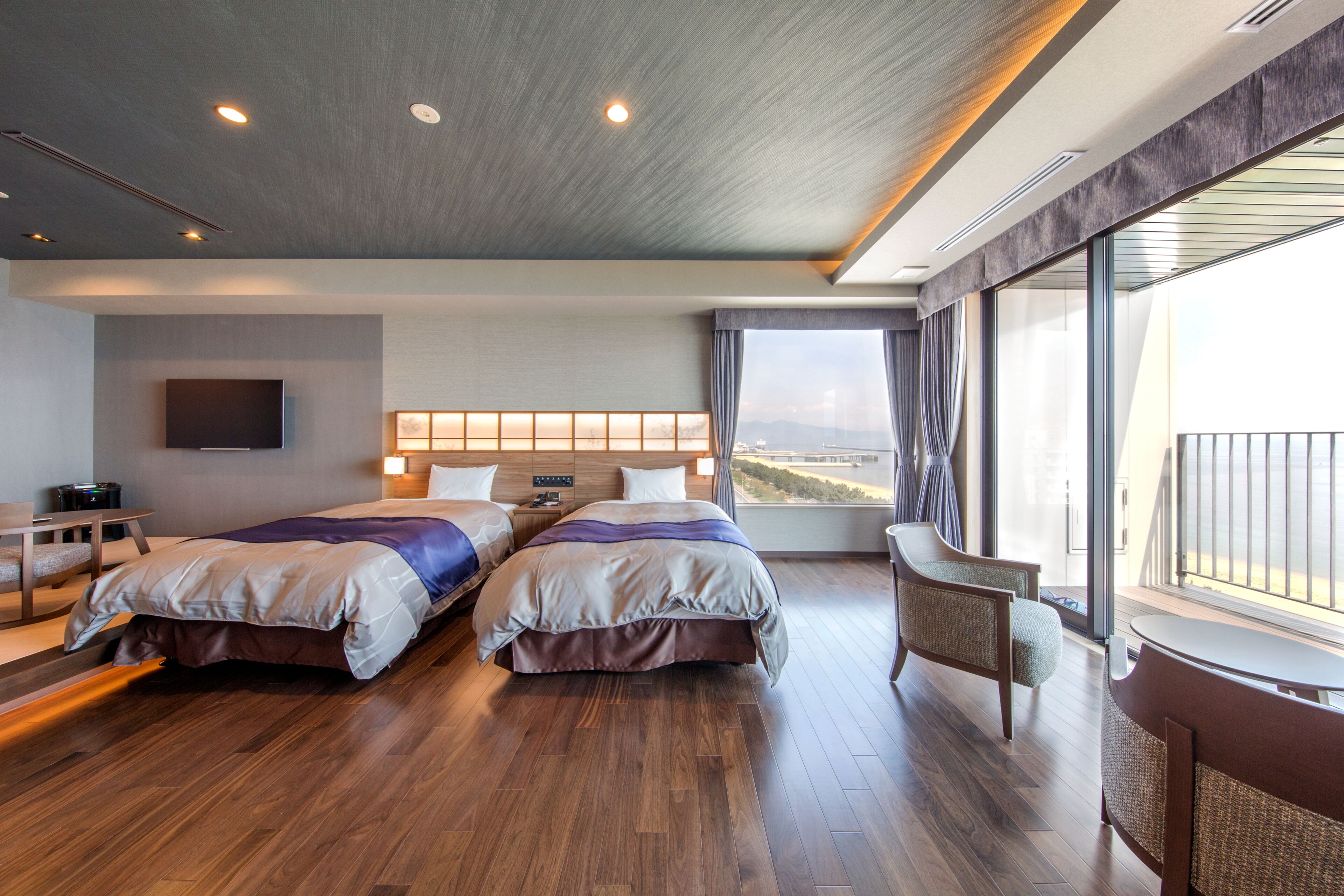 Quarto de hotel com duas camas, uma escrivaninha, uma cadeira e uma janela ampla com vista para a cidade.
