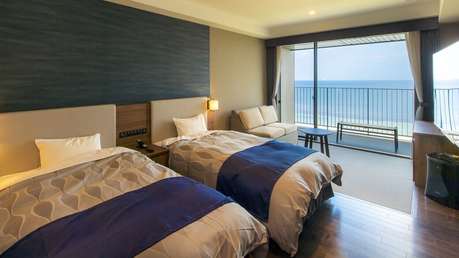 A hotel room with two beds, a balcony with a view, a small table, and a trash bin.