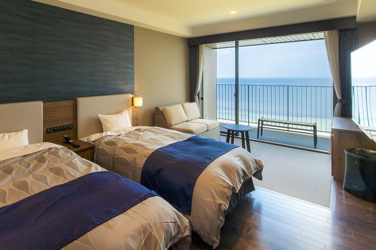 A hotel room with two beds, a balcony with a view, a small table, and a trash bin.